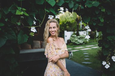 Yazlık elbisesiyle mutlu, genç, sarışın bir kadın. Gülümsüyor ve uzaklara bakıyor. Çiçeklerin yanında yeşil çimlerle birlikte duruyor.