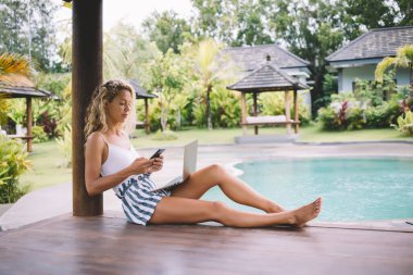 Tüm vücudu, yaz kıyafetleri içinde terasta oturan ve internet üzerinde akıllı telefondan sörf yapan tropikal tatil merkezinden bilgisayarla çalışan odaklanmış genç bir kadın.