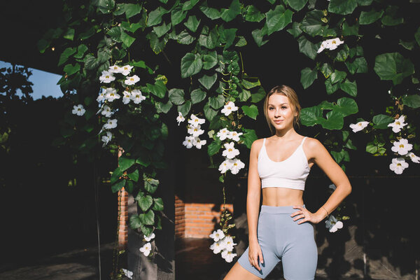 Positive young relaxing female looking away while smiling and standing with hand on waist and bent knee near blooming flowers of park in sunlight during summer vacation