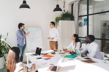 Çok ırklı tasarımcılardan oluşan bir ekip yaratıcı çalışma alanında toplanırken, erkek çalışanlar da çizelgelere işaret eden iş projelerini sunuyorlar.