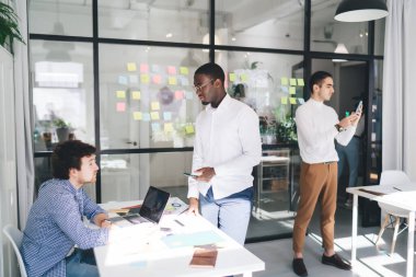 Modern ofisteki proje üzerinde çalışırken, erkek meslektaşı akıllı telefon ekranına bakarken, çok ırklı iş arkadaşlarından oluşan konsantre bir ekip kağıt ve dizüstü bilgisayarla masada toplanıyor.