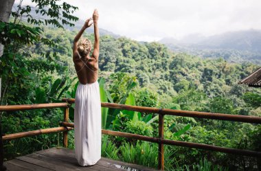 Beyaz elbiseli, kollarını kaldırmış, ahşap terasta duran ve tropikal orman manzarasının keyfini çıkaran tanınmamış kadın gezginin tüm vücudu.