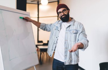 Günlük kıyafetleri ve şapkası içinde gülümseyen yetişkin sakallı adam beyaz tahtanın yanında duruyor ve modern ofiste sunum yaparken grafiği işaret ediyor.