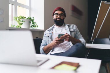 Modern çalışma alanındaki kameraya bakarken, bilgisayarlı masada oturan ve cep telefonunu karıştıran, akıllı kıyafetli, pozitif sakallı, serbest çalışan erkek.