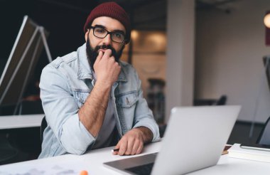 Düşünceli, sakallı, serbest giyinmiş, kırmızı şapkalı, modern çalışma alanındaki kameraya bakan bir hippi.