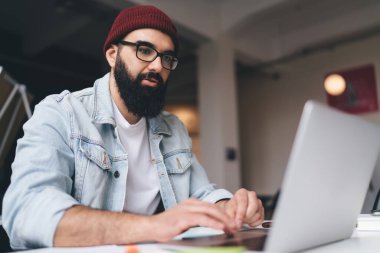 Ciddi sakallı, zeki giysiler ve gözlüklü serbest çalışan, dizüstü bilgisayarlı ve yaratıcı çalışma alanı projesinde çalışan bir erkek.