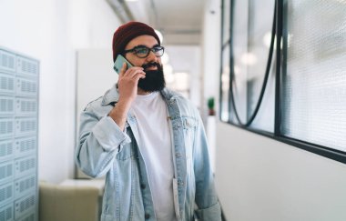 Ofis koridorunda dikilip çalışırken şapkalı ve gözlüklü mutlu bir erkek yönetici cep telefonuyla konuşuyor.