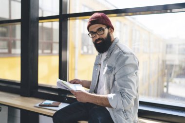 Gözlüklü sakallı bir adamın elinde belgelerle kameraya bakması ve tahta masada oturması, cam pencerenin yanındaki bulanık modern binalara karşı yapışkan notlar.