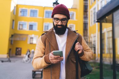 Gözlüklü ve şapkalı şık sakallı hippi güneşli bir günde şehir caddesinde yürürken cep telefonuyla sosyal medyada sohbet ediyor.