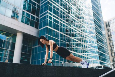 Sınırda şınav çeken siyah aktif giysili, kendine güveni düşük bir sporcu kadın şehir merkezindeki gökdelenlere karşı kararlı görünüyor.