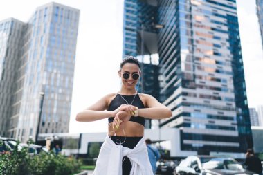 Düşük açılı güneş gözlüklü sporcu kadın kulaklıkla müzik dinlerken ve şehirdeki gökdelenlere karşı dururken takip cihazı kullanıyor.