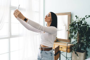 Yandan bakan genç bayan cep telefonuyla selfie çekerken dudakları öpücüklü ve parlak gün ışığında pencerenin yanında duran ekrana bakarken görülüyor.