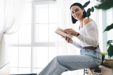 Sıradan kıyafetlerle kitap okuyan, ahşap bir şifoniyerin üzerinde oturup evdeki pencereye bakan genç bir kadının yan görüntüsü.