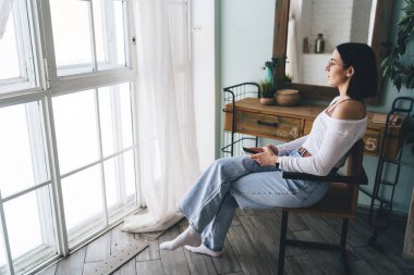 Düşünceli, koyu renk elbiseli, sandalyede oturan, elinde cep telefonuyla pencereden dışarı bakan düşünceli bir kadın.