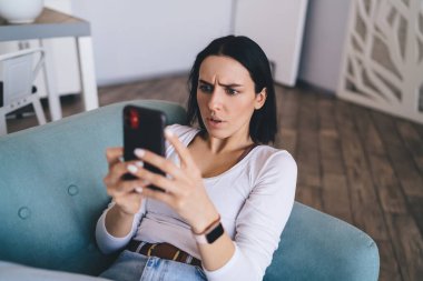 Yüksek açılı, kafası karışık, siyah saçlı, koltukta oturan ve akıllı telefondan şok edici haberler okuyan genç bir kadın.