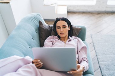 Yukarıdan gelen leylak elbiseli genç kadın rahat koltukta uzanıyor ve evdeki güneşli oturma odasında netbook kullanıyor.