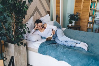 Yüksek açılı, rahat giyinen, kitap okuyan ve yatak odasında uzanan genç bir kadın.