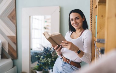 Hafta sonu evde, kitaplığın yanında kahverengi kapaklı ilginç bir kitapla gündelik giysiler içinde mutlu genç bayan kitap kurdu duruyor.