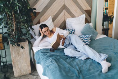 Yatakta uzanmış, yatak odasında dinlenirken kitap okuyan, gündelik giysiler içinde tam vücut açılı genç bir kadın.