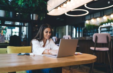 Bilgisayara bakan, gülümseyen ve ahşap masada akıllı telefon işaretleyici notlarıyla oturan genç, pozitif serbest çalışan bir bayan kafe işletme projesinde çalışıyor.