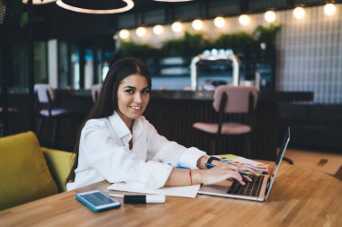 Akıllı yüzlü, sırıtan genç bir serbest çalışan dizüstü bilgisayarlı akıllı telefon işaretleme belgeleriyle masada oturuyor ve kafedeki modern çalışma alanında çalışırken kameraya bakıyor.