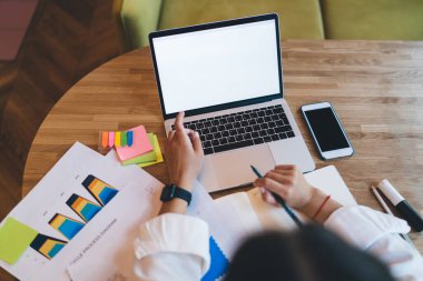 Beyaz bluzlu isimsiz kadın serbest yazar, tahta masada boş ekranlı dizüstü bilgisayarla oturuyor ve proje sırasında defterine notlar alıyor.