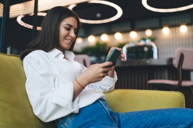 Pozitif genç bayan gülümseyip göz gezdirirken ekrandaki cep telefonuna bakıyor ve molada bulanık kafeye karşı rahat koltukta oturuyor.