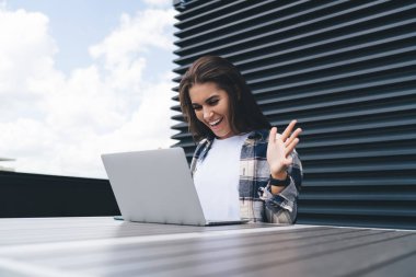 Neşeli, genç, serbest çalışan, gündelik giysiler içinde ellerini kaldırarak masada oturan ve terasta yaptığı çalışmalar sırasında sevinen genç bir bayandan.