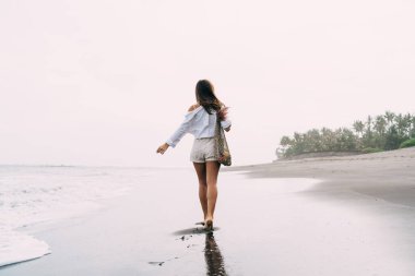 Tişörtlü ve şortlu genç bir kadının çıplak ayakla çıplak ayakla kumsalda yürüdüğünü kolları açık bir şekilde dalgalanan deniz suyunun yanında.