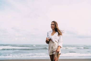 Kumlu deniz kıyısında kitap ve ip torbasıyla dikilen mutlu bir kadın.