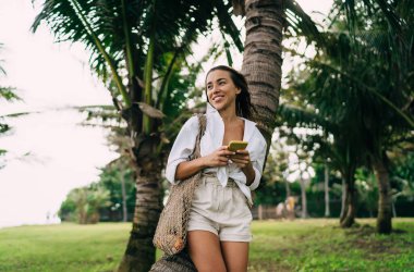 Çevre dostu çantalı neşeli bir kadın yeşil tropikal bir parkta dururken ve elinde cep telefonuyla elinde cep telefonuyla dikilirken gülümsüyor ve uzaklara bakıyor.