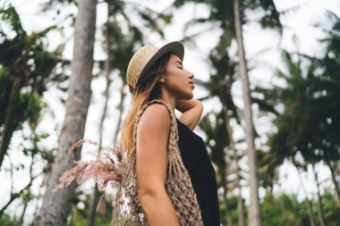 El çantalı ve şapkalı genç bayanın düşük açılı manzarası yeşil parkta gözleri kapalı dururken güneşli bir günün tadını çıkarırken bulanık parktaki palmiye ağaçlarının arasında.