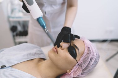 Güzellik kliniğinde gözlüklü etnik hastaya lazerle yüz kılı çıkarma işlemi yapan profesyonel kadın güzellik uzmanının yan görüntüsü