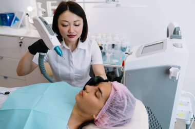 Çağdaş kuaförde güzellik prosedürü sırasında kadın müşterilerin yüzüne YAG lazeri kullanarak siyah eldivenli profesyonel kozmetikçi.