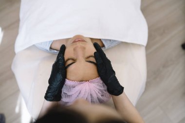 Kadın müşterinin yüksek açılı gözleri kapalı bir şekilde masada yatarken siyah eldiven masajı yapan isimsiz bir uzman kesiliyor.