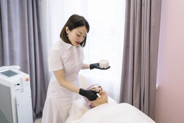 Eldivenli ve üniformalı genç Asyalı kadın kozmetik uzmanının ameliyat sırasında müşterinin yüzüne sıvı maske takması.