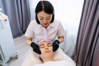 Üniformalı genç kozmetikçinin, güzellik kliniğinde cilt bakımı sırasında kadın müşterilerin yüzüne nemlendirici krem sürmesinden.