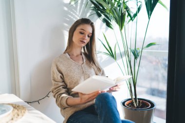 İlgili bayan öğrenci günlük giysiler giyip ders kitabı okuyor ve pencerenin yanında oturup uzaktan öğrenirken ev ödevlerini yapıyor.