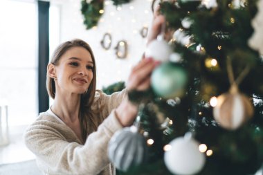 Yılbaşı partisine hazırlanırken, mutlu, genç bir bayanın gülümsemesi ve takılarını Noel ağacına asması