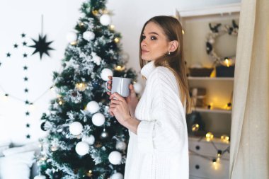 Süslü bir odada sıcak bir bardak sıcak içecekle dikilen ve Noel ağacıyla süslenmiş genç bir kadının yan görüntüsü.