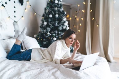 Sıcak giysiler içinde, gözlük takan, dizüstü bilgisayarlı dokunmatik bilgisayarla program testi yapan ve Noel ağacının yanındaki ekrana bakan yoğun genç kadın serbest yazar.