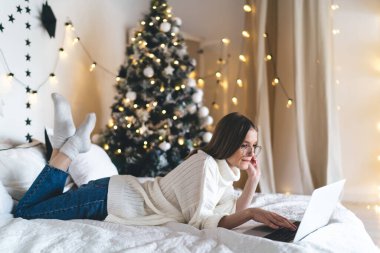 Yatakta yatıp dizüstü bilgisayarla çalışan genç kadın yazılım test edicisinin yan görüntüsü ve Noel ağacı olan dekore edilmiş bir odada uzaktan kumandalı şirket için yapılan testler.