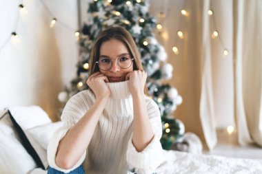 Sıcak beyaz süveter ve gözlüklü genç bir kadın Noel için yatak odasında yatakta dinleniyor ve kameraya bakıyor.