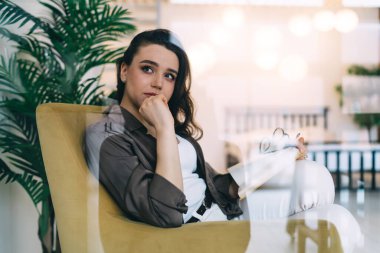 Düşünceli genç bayanlardan oluşan camdan bir duvarın arkasına bakın. Düşünceli, rahat giysiler içinde, saksı bitkisinin yanındaki rahat koltukta dinleniyor. Düşünceli bir şekilde başka tarafa bakıyor.