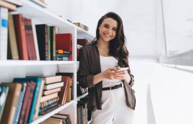 Kitaplığın yanında duran ve beyaz geniş kütüphane odasındaki kameraya bakarken akıllı telefon kullanan gündelik kıyafetlerle uzun siyah dalgalı saçlı pozitif genç bir kadın.