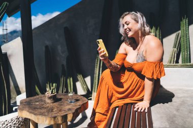 Neşeli genç bayan cep telefonuyla ahşap masada oturuyor ve ekrana bakıyor ve gülümsüyor mesaj okuyor ve kafede bir bardak suyla gün ışığında dinleniyor.