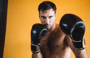 Spor salonunda spor yaparken siyah kum torbasına yakın duran ve kameraya bakan güçlü üstsüz, kaslı erkek boksör.