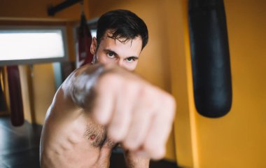 Spor salonunda egzersiz yaparken kameraya bakarken çıplak elleriyle havaya yumruk atan kararlı, üstsüz bir adam.