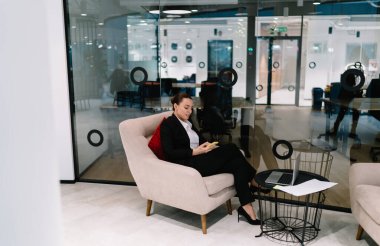 Resmi kıyafetli genç bayan çalışan, modern yaratıcı ofiste dizüstü bilgisayar ve cam duvar kullanarak koltukta oturuyor.