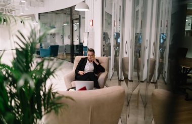 Resmi kıyafetler içinde genç bir girişimci rahat koltukta oturuyor ve yaratıcı çalışma alanındaki cam duvarların yanında cep telefonuyla konuşuyor.